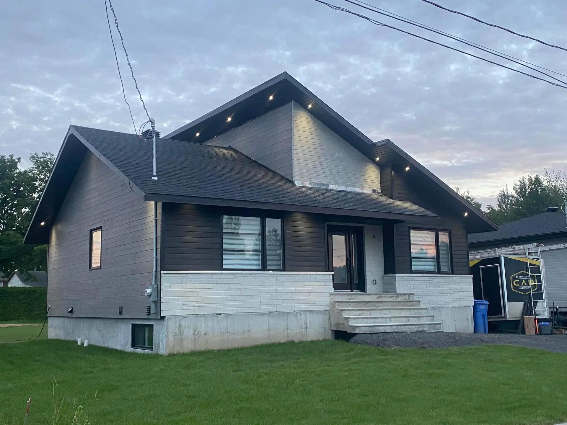 A modern house with dark wood siding and white stone on the lower facade. It has a gable roof with recessed lights under the eaves, a concrete staircase leading to the main entrance, and a garage visible on the right. The lawn is well-maintained, and the sky is slightly overcast at dusk.