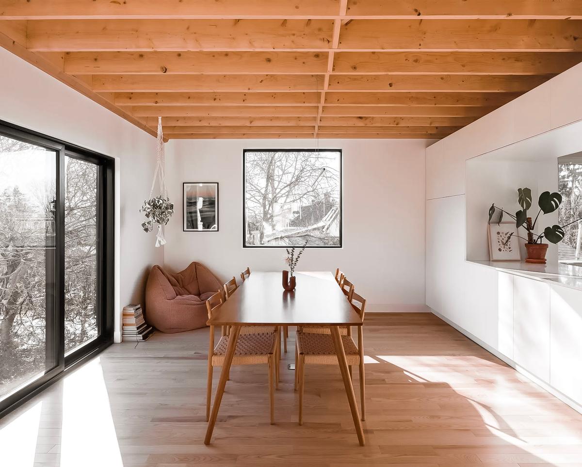 Bright dining room with a wooden table and chairs, exposed beam ceiling, large windows, and minimalist decor.