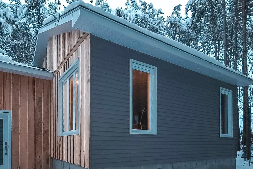 Home addition with a wooden facade, modern windows, and concrete foundation surrounded by a snowy forest.