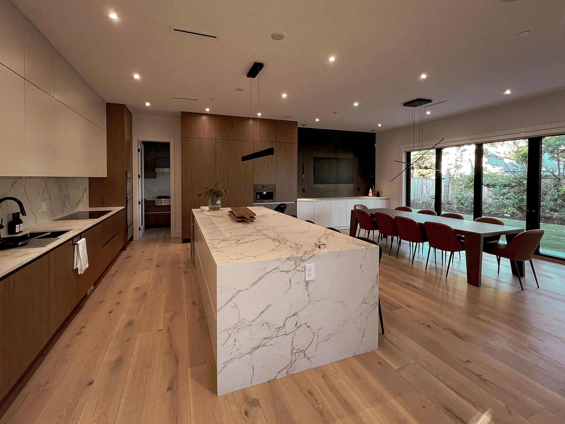 Open modern kitchen with white veined marble island, wooden cabinets, light wood flooring, large dining table with red chairs, and designer pendant lighting.