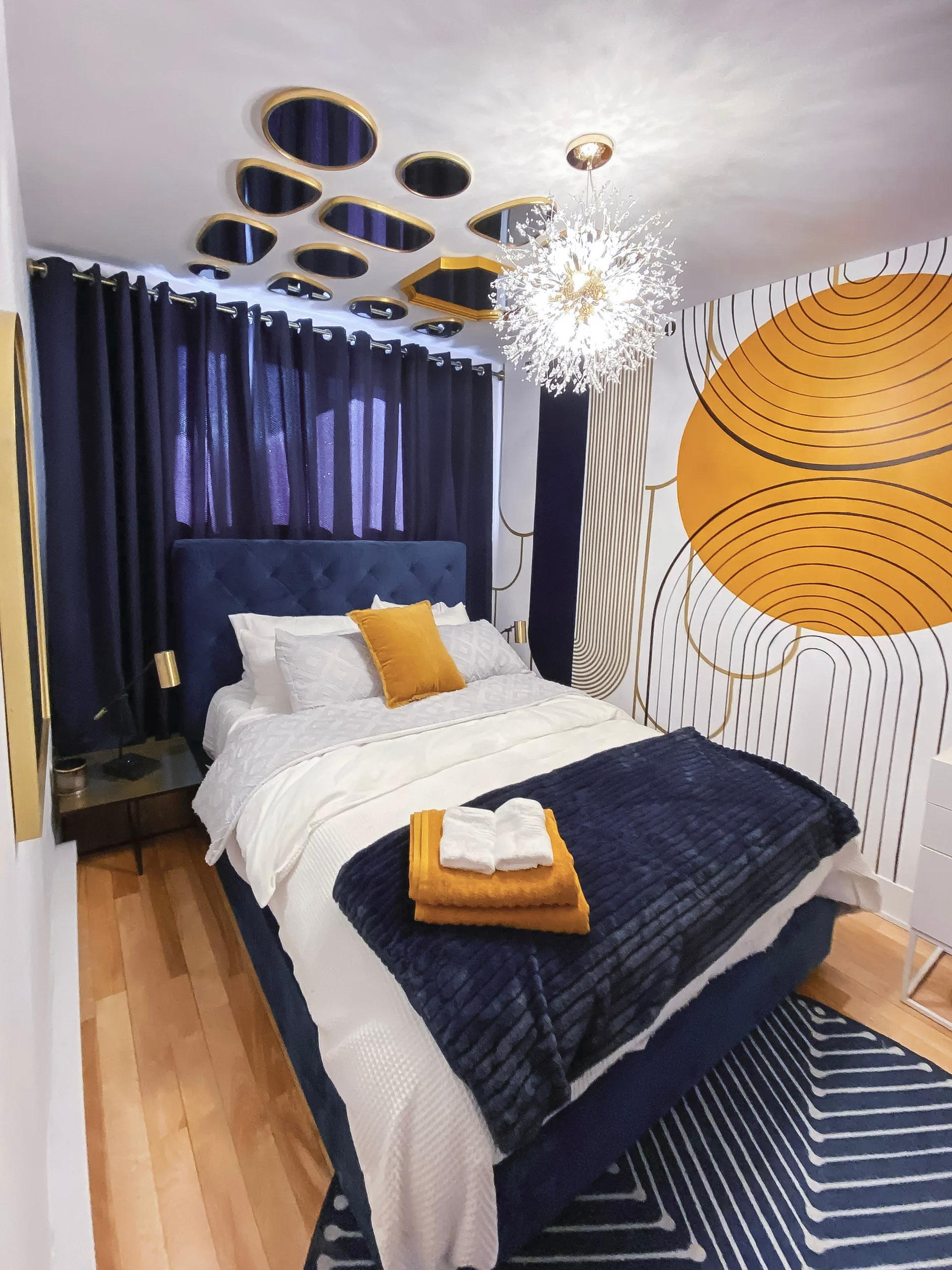 Modern bedroom with blue tufted bed, graphic yellow and white wall decor, designer ceiling light, and dark curtains for a warm and elegant atmosphere.