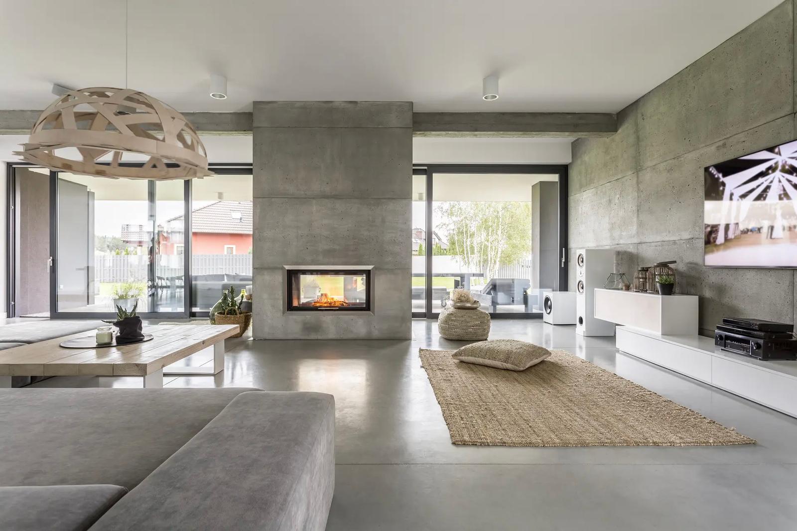 Modern living room with an industrial style featuring raw concrete walls, polished concrete flooring, a built-in fireplace, and large glass doors opening to an outdoor terrace.