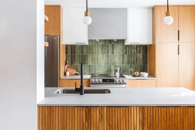 Scandinavian-looking Montréal kitchen, featuring a wood-panelled central island. The light wooden cabinets and green ceramic tiles make for a harmonious contrast. The quartz countertop and light wooden flooring brighten up the space.