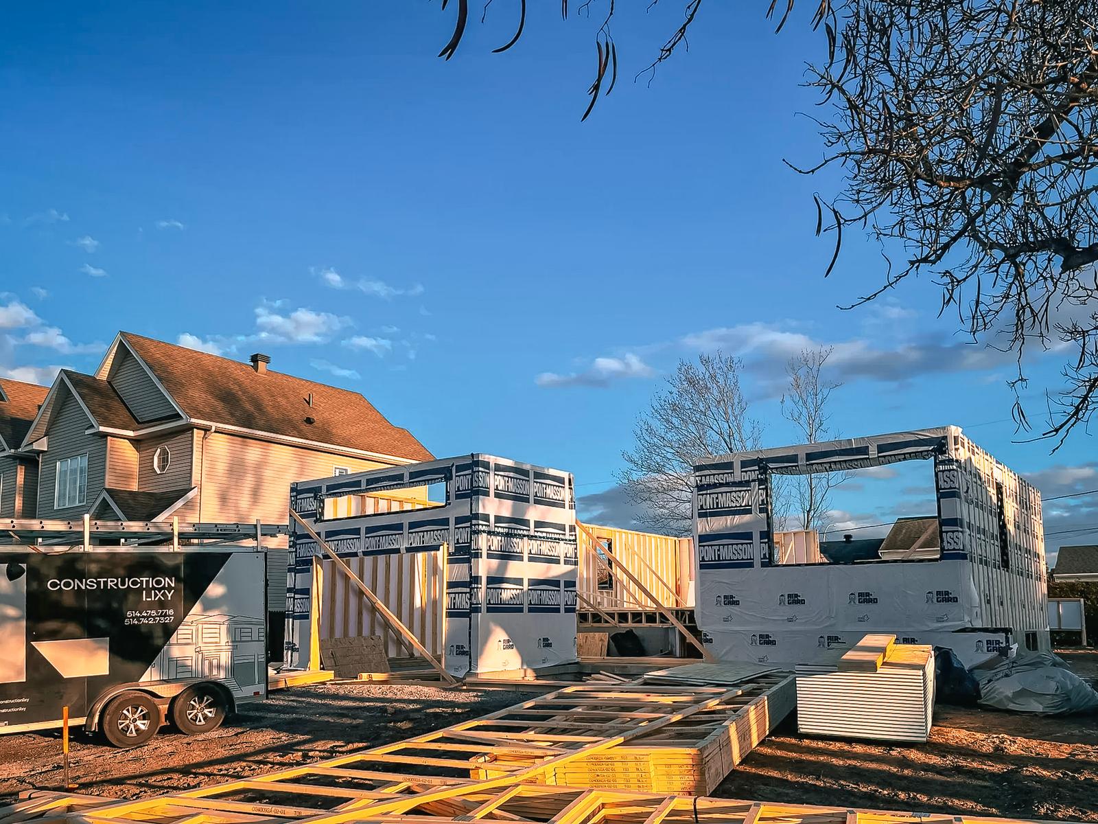 Construction of a wooden frame house with stacked materials and a construction trailer at sunset.<br>