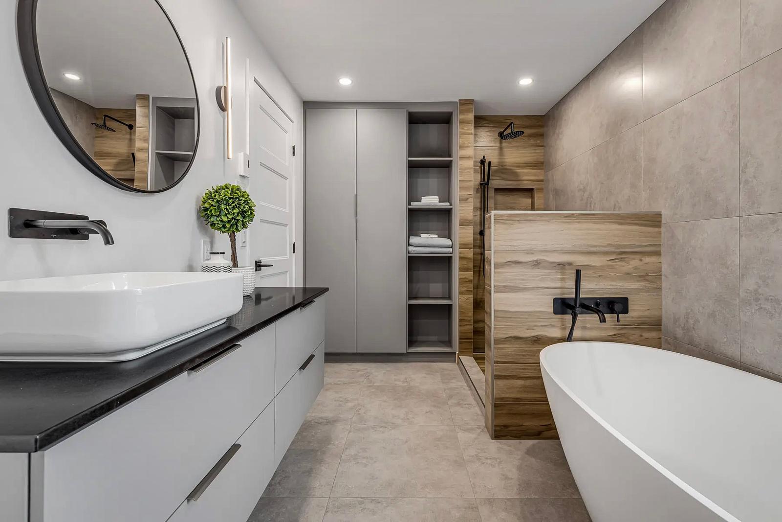 Modern bathroom with freestanding bathtub, open shower with wood finish, and gray tiled walls. Gray vanity with black countertop, white vessel sink, and matte black fixtures.