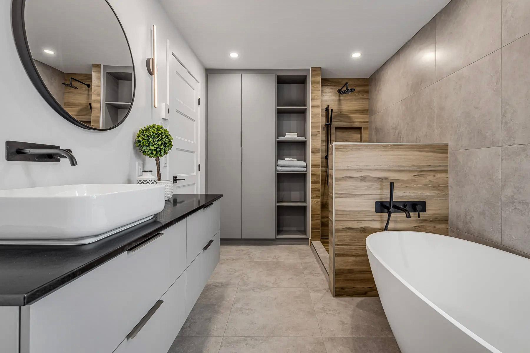 Modern bathroom with freestanding bathtub, open shower with wood finish, and gray tiled walls. Gray vanity with black countertop, white vessel sink, and matte black fixtures.