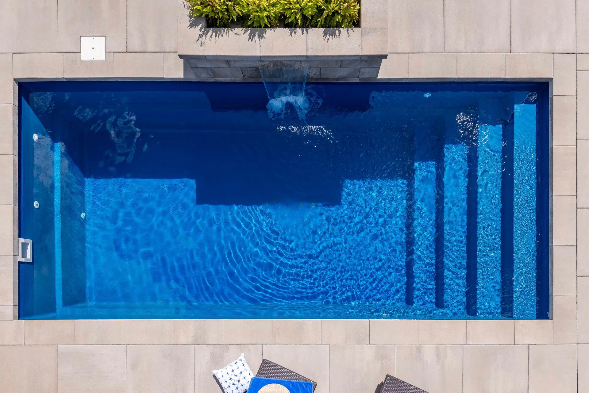 Aerial view of a rectangular swimming pool with an integrated waterfall, surrounded by modern tiles and decorative vegetation.