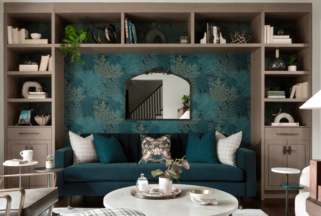 Modern living room with a blue velvet sofa, floral wallpaper, designer mirror, built-in bookshelf, and marble coffee table in a bright and elegant space.