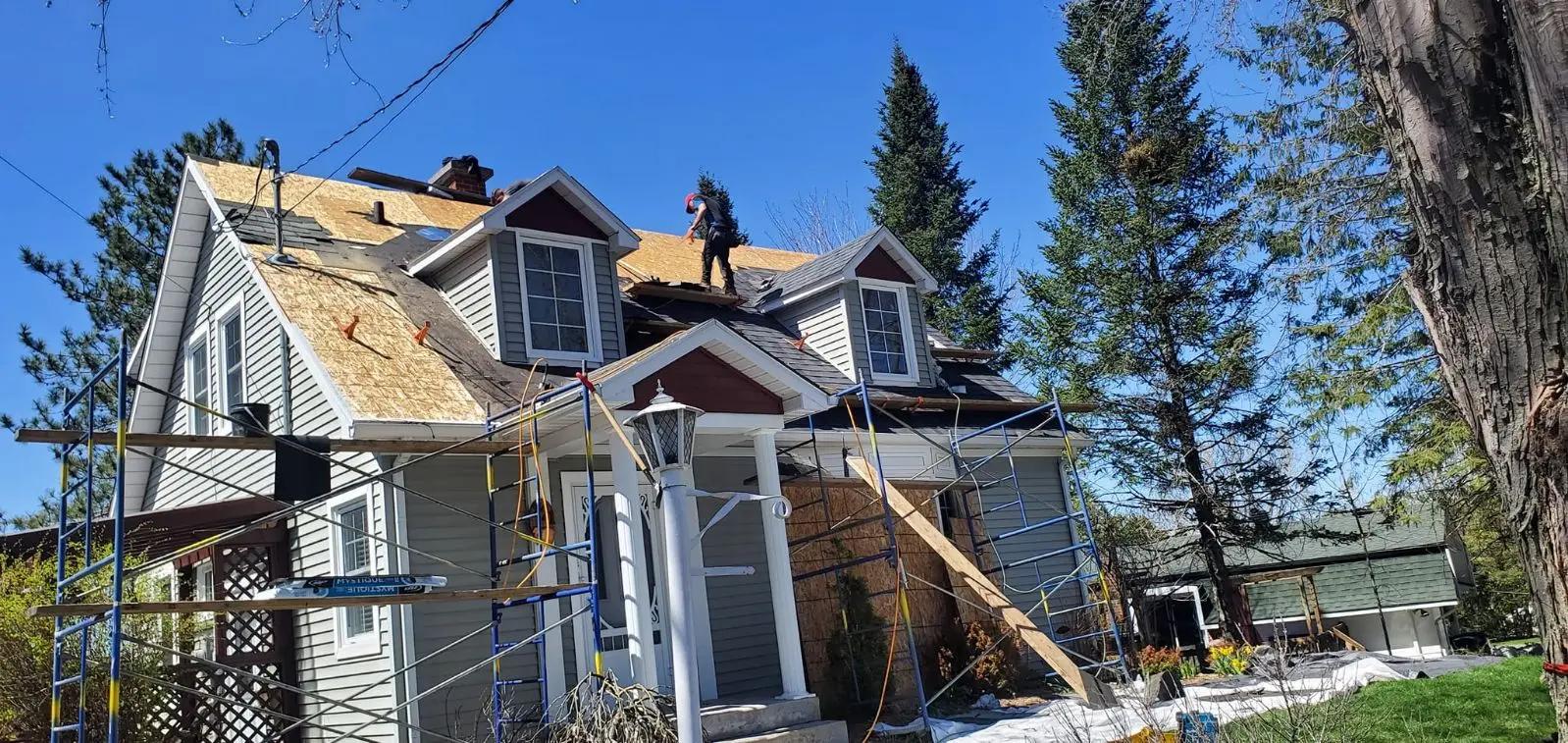 Roof renovation work on a two-story residential house. Two workers are on the roof installing plywood panels, surrounded by scaffolding for safety. The house, set in a green, tree-filled environment, features a gray facade with white dormers and a covered front porch